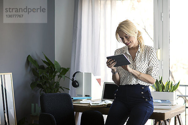 Lächelnde Frau  die einen Tablet-Computer benutzt  während sie sich im Heimbüro an den Tisch lehnt