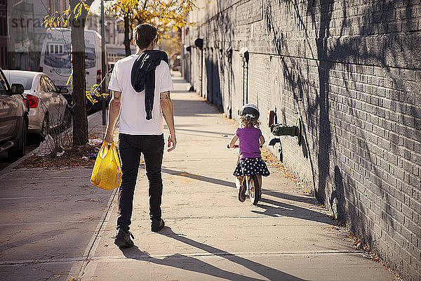 Rückansicht eines Vaters  der zu Fuß geht  während die Tochter auf dem Fußweg radelt