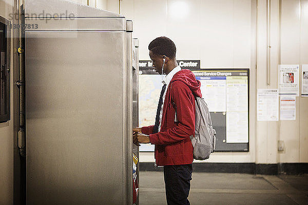 Seitenansicht eines Teenagers in Schuluniform am Fahrkartenautomaten am Bahnhof