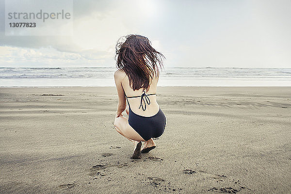 Rückansicht einer am Bethells Beach kauernden Frau gegen Meer und Himmel