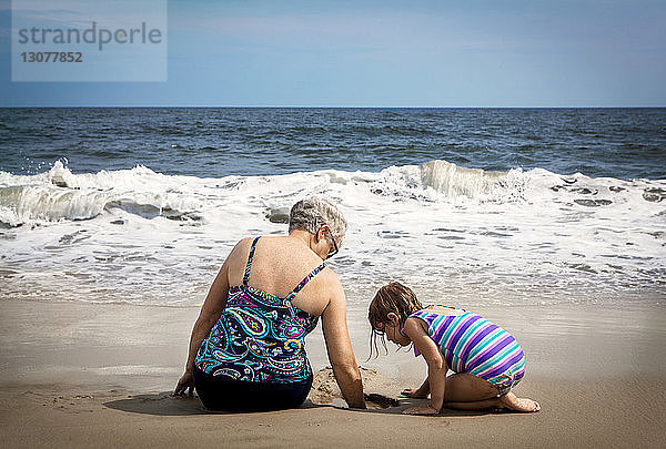Mädchen schaut nach unten  während die Großmutter am Strand im Sand buddelt