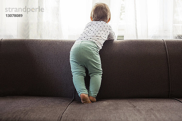 Rückansicht eines kleinen Mädchens  das durch das Fenster schaut  während es zu Hause auf dem Sofa steht