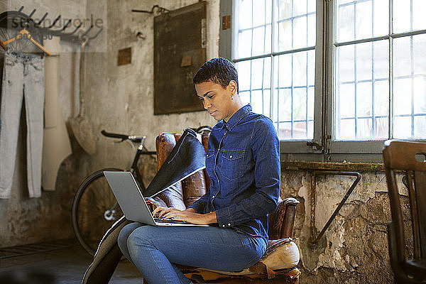 Seriöse Frau benutzt Laptop  während sie in alter Werkstatt auf einem Stuhl sitzt
