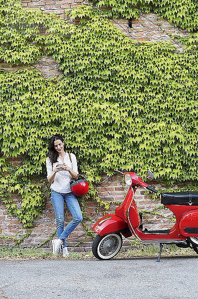 Frau benutzt Mobiltelefon  während sie auf einem Motorroller an einer Efeuwand steht