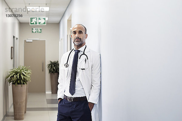 Porträt eines Arztes mit an die Wand gelehnten Händen in Taschen im Krankenhaus