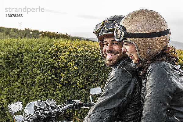 Fröhliches Paar unterhält sich im Sitzen auf Motorrad gegen Himmel
