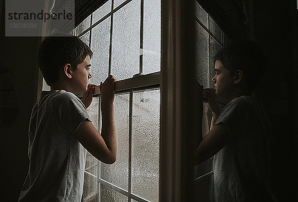 Seitenansicht eines nachdenklichen Jungen  der durch ein nasses Fenster schaut und sich zu Hause im Spiegel spiegelt