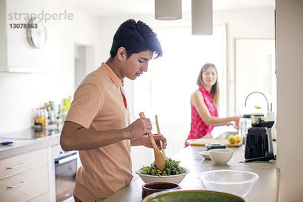 Seitenansicht eines Mannes bei der Salatzubereitung mit einer im Hintergrund stehenden Frau in der Küche