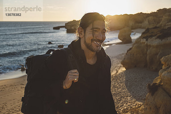 Lächelnder Mann mit Rucksack steht bei Sonnenuntergang am Strand gegen den Himmel