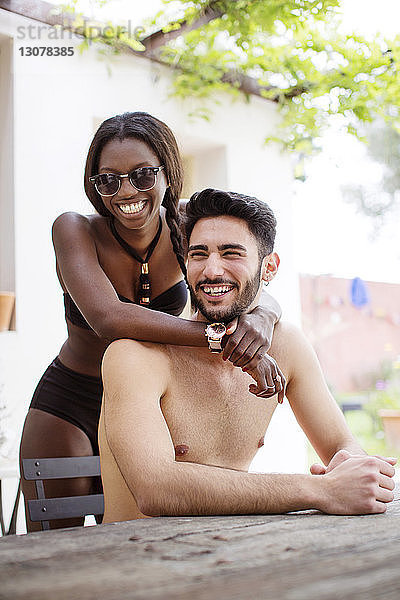 Porträt einer glücklichen Frau im Bikini  die mit Armen um den hemdlosen Mann im Hof steht