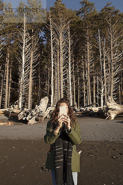 Junge Frau fotografiert mit einem Smartphone vor Bäumen am Strand