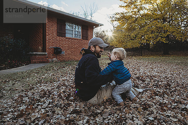 Glücklicher Vater spielt mit seinem Sohn  während er auf Herbstlaub im Hof sitzt
