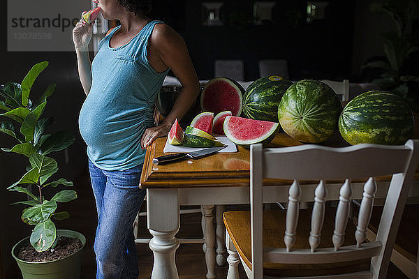 Mittelsektion einer schwangeren Frau  die am Tisch steht und Wassermelone isst