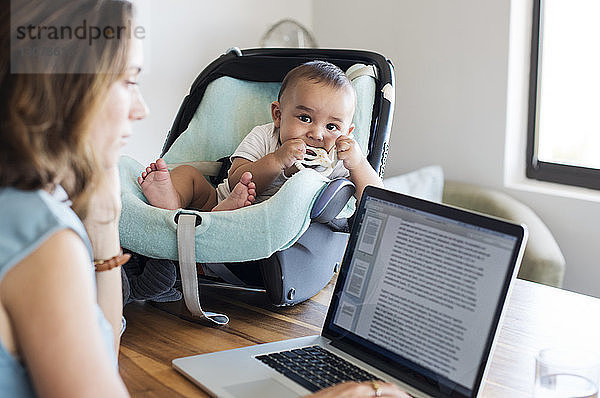 Mutter benutzt Laptop-Computer von einem kleinen Jungen im Kindersitz auf einem Tisch zu Hause