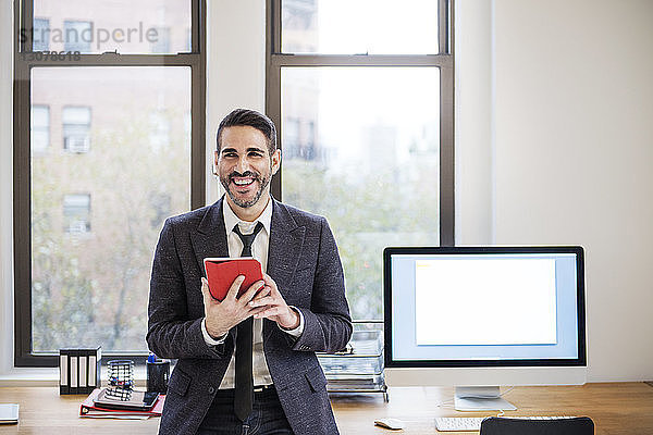 Glücklicher Geschäftsmann  der einen Tablet-Computer benutzt  während er sich im Büro auf den Schreibtisch lehnt
