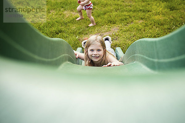 Porträt eines glücklichen Mädchens  das rutscht  während der Bruder auf dem Spielplatz spielt