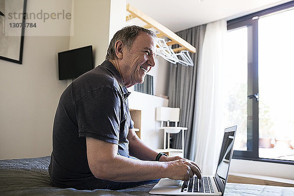 Seitenansicht eines fröhlichen älteren Mannes  der weg schaut  während er den Laptop im Schlafzimmer benutzt