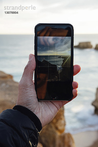 Abgetrennte Hand eines Mannes  der bei Sonnenuntergang das Meer mit einem Mobiltelefon fotografiert