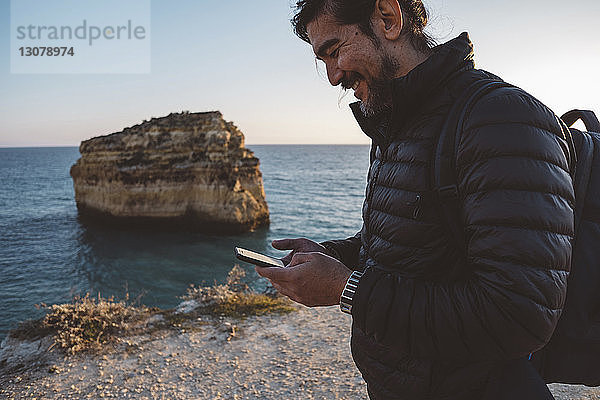 Seitenansicht eines Mannes  der ein Smartphone benutzt  während er bei Sonnenuntergang am Strand vor klarem Himmel steht