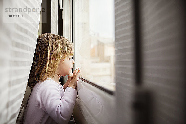 Seitenansicht eines neugierigen Mädchens  das zu Hause durch ein Fenster schaut