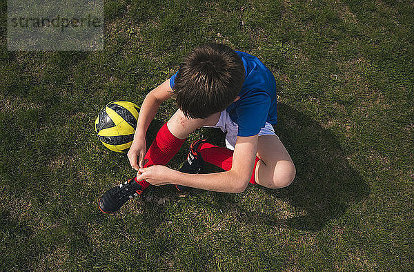 Schrägansicht eines Jungen beim Schnürsenkelbinden  der auf einem Fussballfeld sitzt