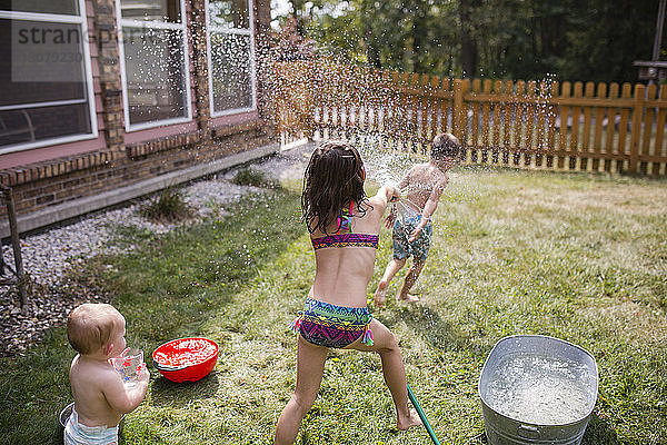 Kleines Mädchen sieht Schwester an  die durch einen Schlauch Wasser auf hemdlosen Bruder spritzt