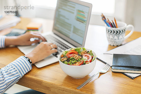 Beschnittenes Bild einer Geschäftsfrau  die am Laptop-Computer an einer Salatschüssel auf einem Holztisch im Heimbüro bloggt