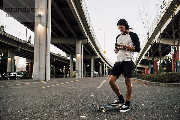 Mann benutzt Mobiltelefon  während er mit Skateboard auf der Straße steht