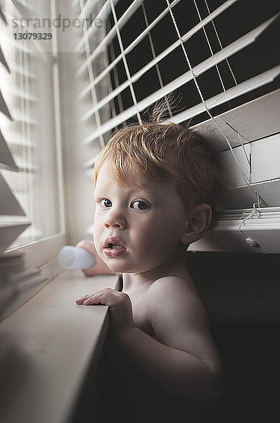 Porträt eines hemdlosen kleinen Jungen  der zu Hause am Fenster steht