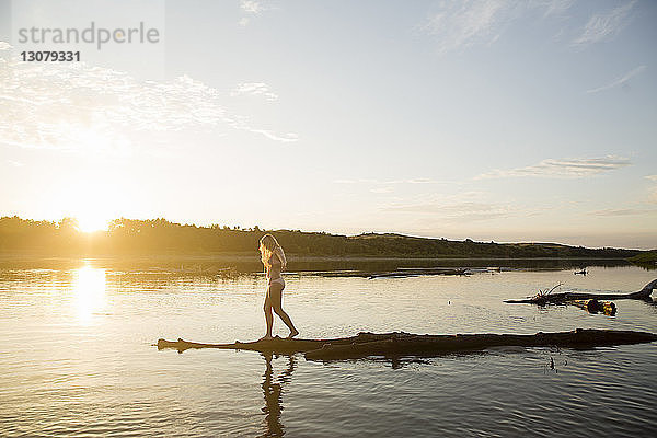 Seitenansicht einer jungen Frau im Bikini  die bei Sonnenuntergang am Flussufer gegen den Himmel steht