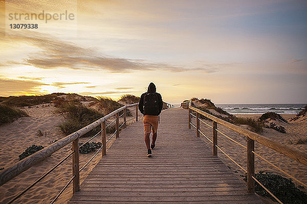 Rückansicht eines Wanderers  der bei Sonnenuntergang auf einem Steg am Strand gegen den Himmel läuft