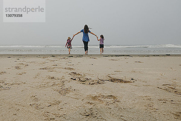 Rückansicht einer Mutter  die die Hände ihrer Töchter hält  während sie am Strand gegen den Himmel steht