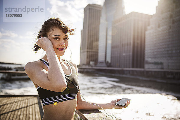 Porträt einer lächelnden Frau  die mit einem Smartphone am Fluss Musik hört