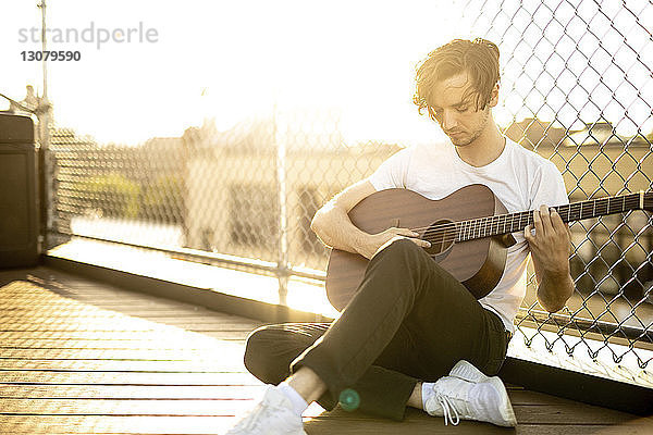 Mann spielt Gitarre  während er bei Sonnenuntergang am Maschendrahtzaun gegen den Himmel sitzt