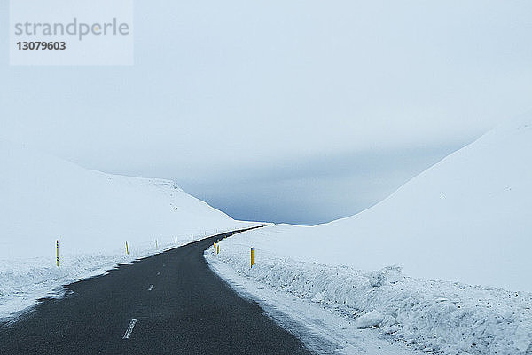Leere Landstraße inmitten eines verschneiten Feldes vor bewölktem Himmel