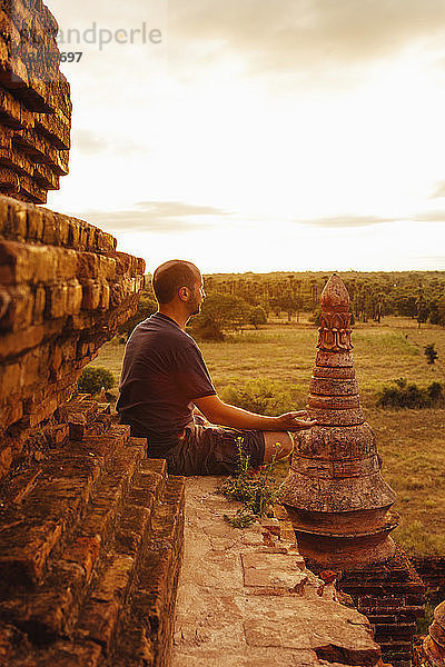 Seitenansicht eines Menschen  der meditiert  während er bei Sonnenuntergang auf einem buddhistischen Tempel am Himmel sitzt