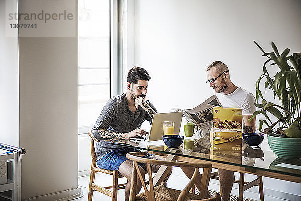 Homosexuelles Paar benutzt Laptop und liest Zeitung am Frühstückstisch