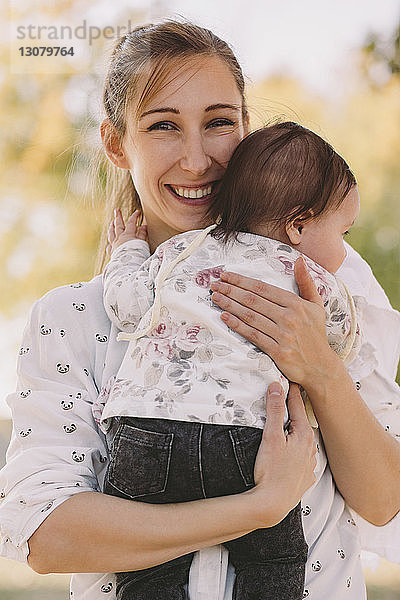 Porträt einer glücklichen Mutter  die eine Tochter trägt  während sie im Herbst im Park steht