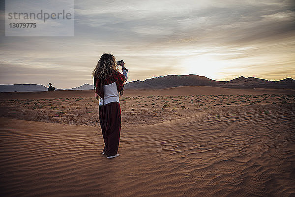 Rückansicht einer Frau  die durch ein Mobiltelefon in der Wüste vor bewölktem Himmel fotografiert