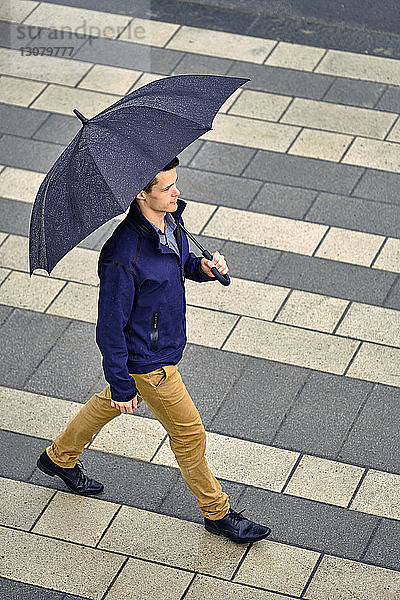 Hochwinkelaufnahme eines jungen Geschäftsmannes mit Regenschirm  der bei Regen auf dem Bürgersteig in der Stadt geht