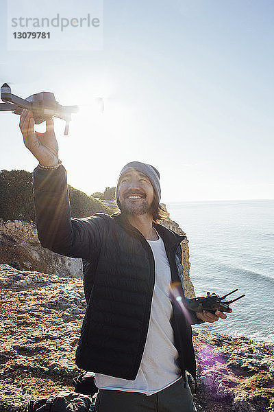 Lächelnder Mann fliegt Quadcopter  während er am Strand gegen den klaren Himmel am sonnigen Tag steht