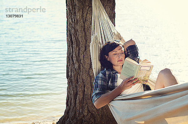 Frau liest Buch  während sie sich in der Hängematte am Seeufer am Baum entspannt