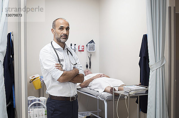 Porträt eines selbstbewussten Arztes  der im medizinischen Untersuchungsraum mit dem Patienten im Hintergrund steht