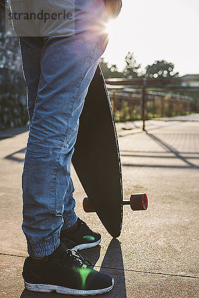Niedriger Teil eines Mannes  der ein Skateboard hält  während er bei Sonnenuntergang gegen den Himmel steht