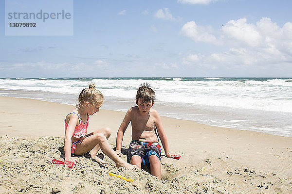 Geschwister spielen mit Sand am Strand gegen den Himmel während eines sonnigen Tages