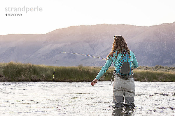 Rückansicht einer jungen Frau mit Rucksack beim Fliegenfischen im Owens River vor Bergen