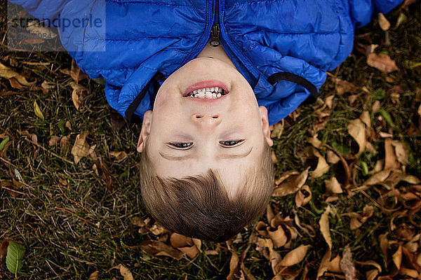 Kopfporträt eines fröhlichen Jungen  der im Herbst im Park auf trockenem Laub liegt