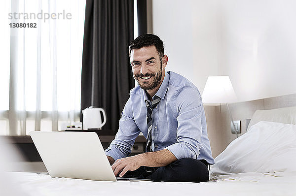 Porträt eines glücklichen Geschäftsmannes  der mit Laptop auf dem Bett im Hotelzimmer sitzt