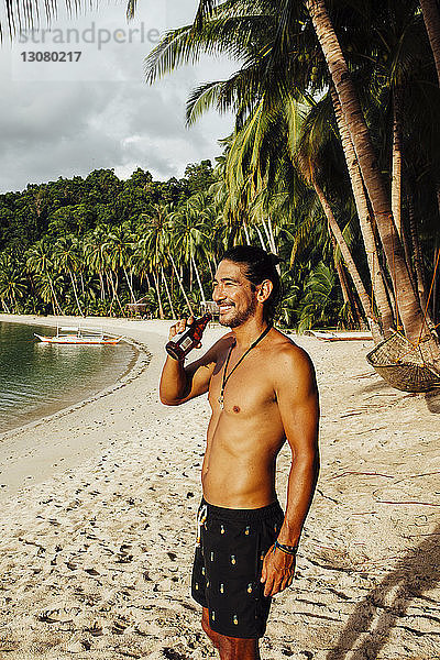 Fröhlicher Mann ohne Hemd trinkt am Strand stehend