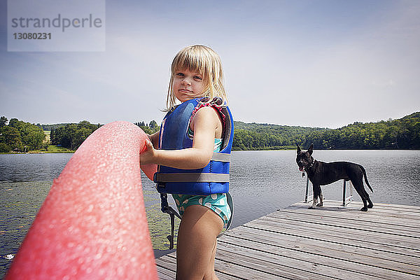 Porträt eines Mädchens  das ein Nudelschwimmen hält  während es mit einem Hund auf dem Pier steht